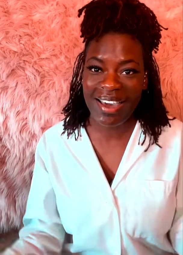 Raven Baxter, in a white lab coat with her locs pulled up, smiles mid-line in front of a pink faux-fur backdrop.