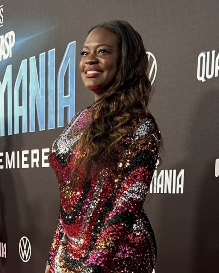 Dr. Raven Baxter smiles in a long-sleeved sequined dress striped in pink, red, silver and black, her long hair over one shoulder, in front of a dark film premiere backdrop.