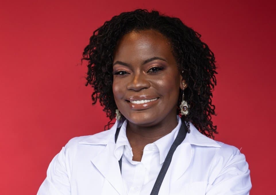 Raven Baxter smiles in a white lab coat with a dark lanyard in front of a red wall.