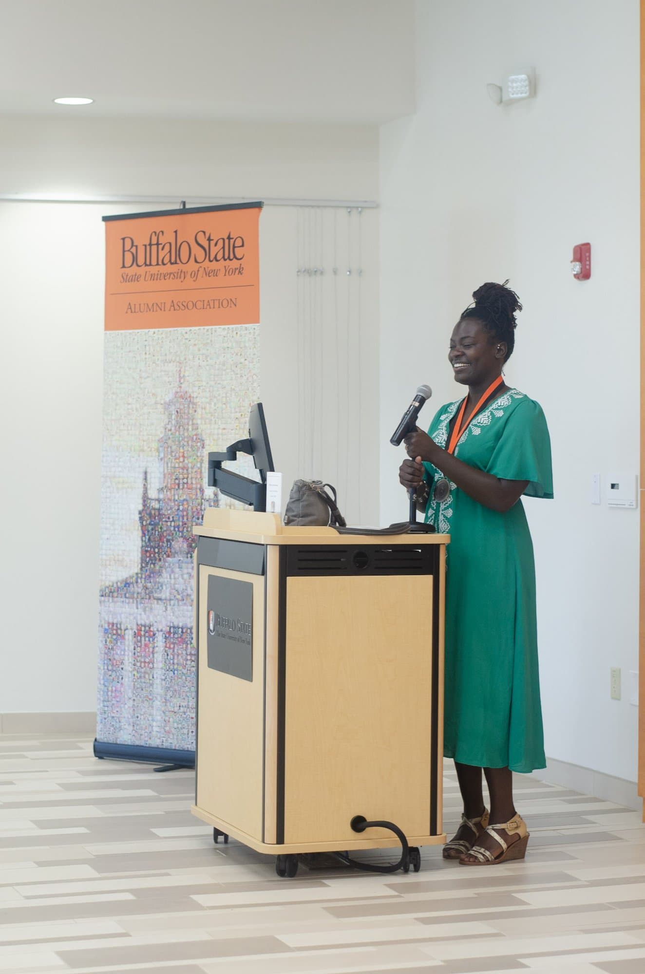 Raven Baxter, in a green embroidered dress and a medal on an orange ribbon, smiles as she holds a microphone beside a lectern and a tall Buffalo State Alumni Association banner.