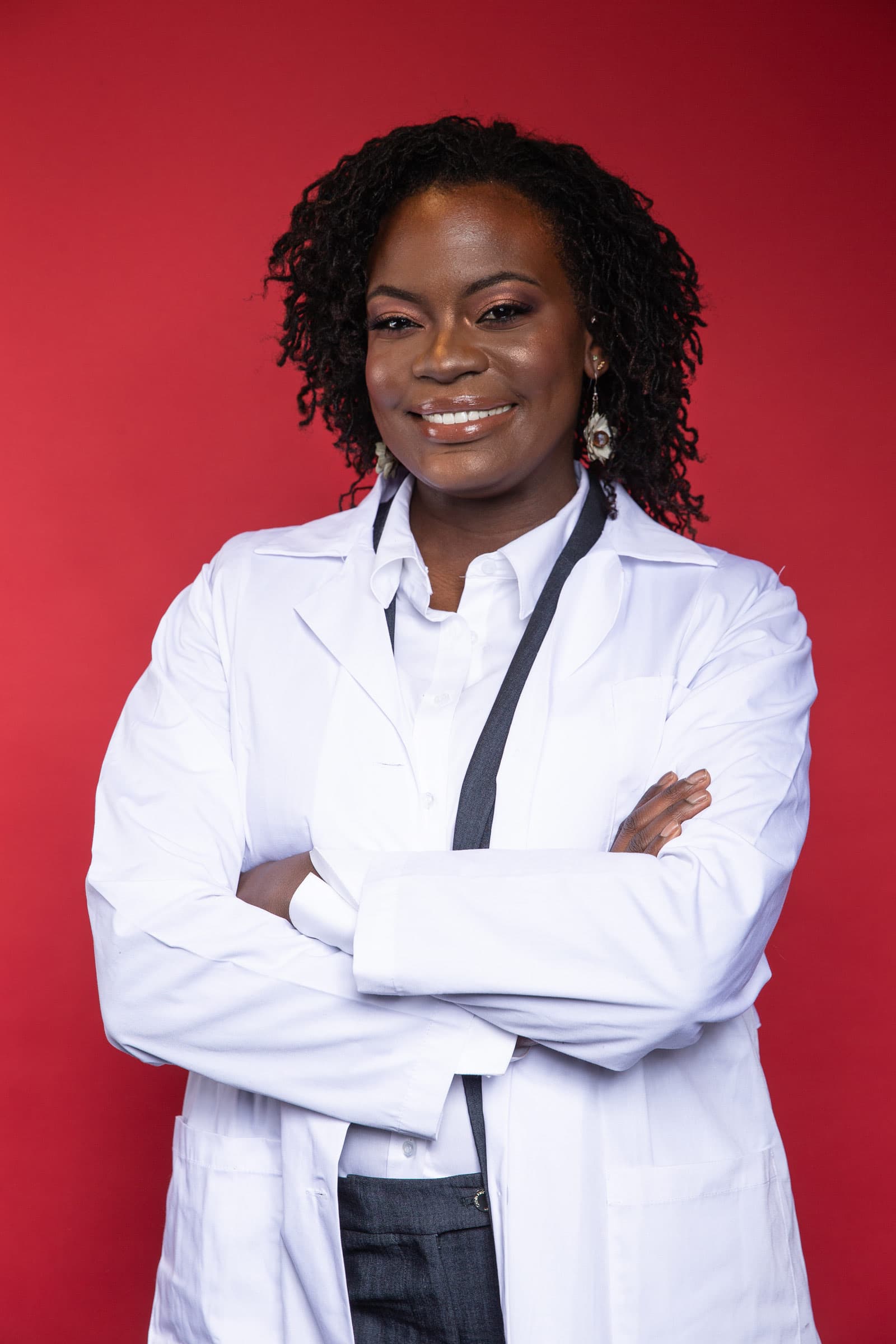 Raven Baxter smiles with her arms crossed, wearing a white lab coat and a dark lanyard, in front of a red wall.