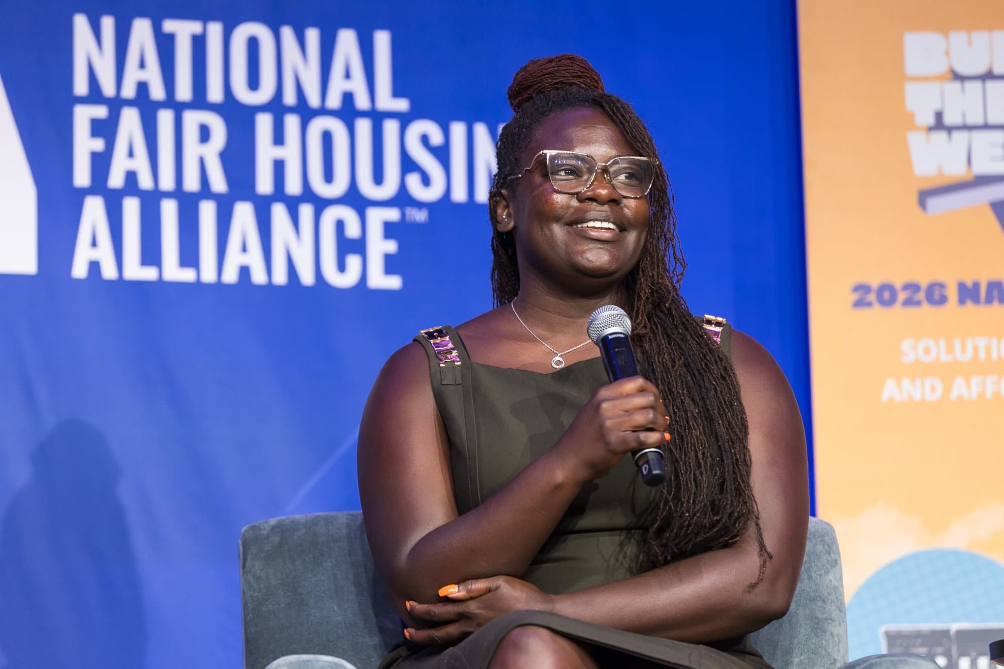 Dr. Raven Baxter, in glasses and an olive dress, smiles as she holds a microphone in front of a blue National Fair Housing Alliance backdrop.