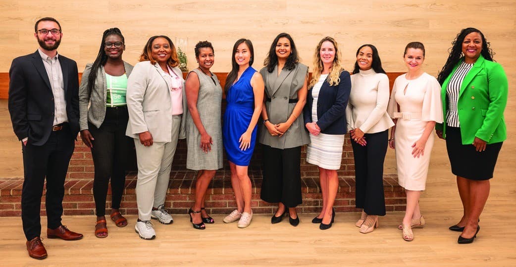 Ten people stand in a row in front of a light wood wall and a brick ledge. Baxter, second from left, wears braids, glasses, a gray blazer and a green T-shirt.