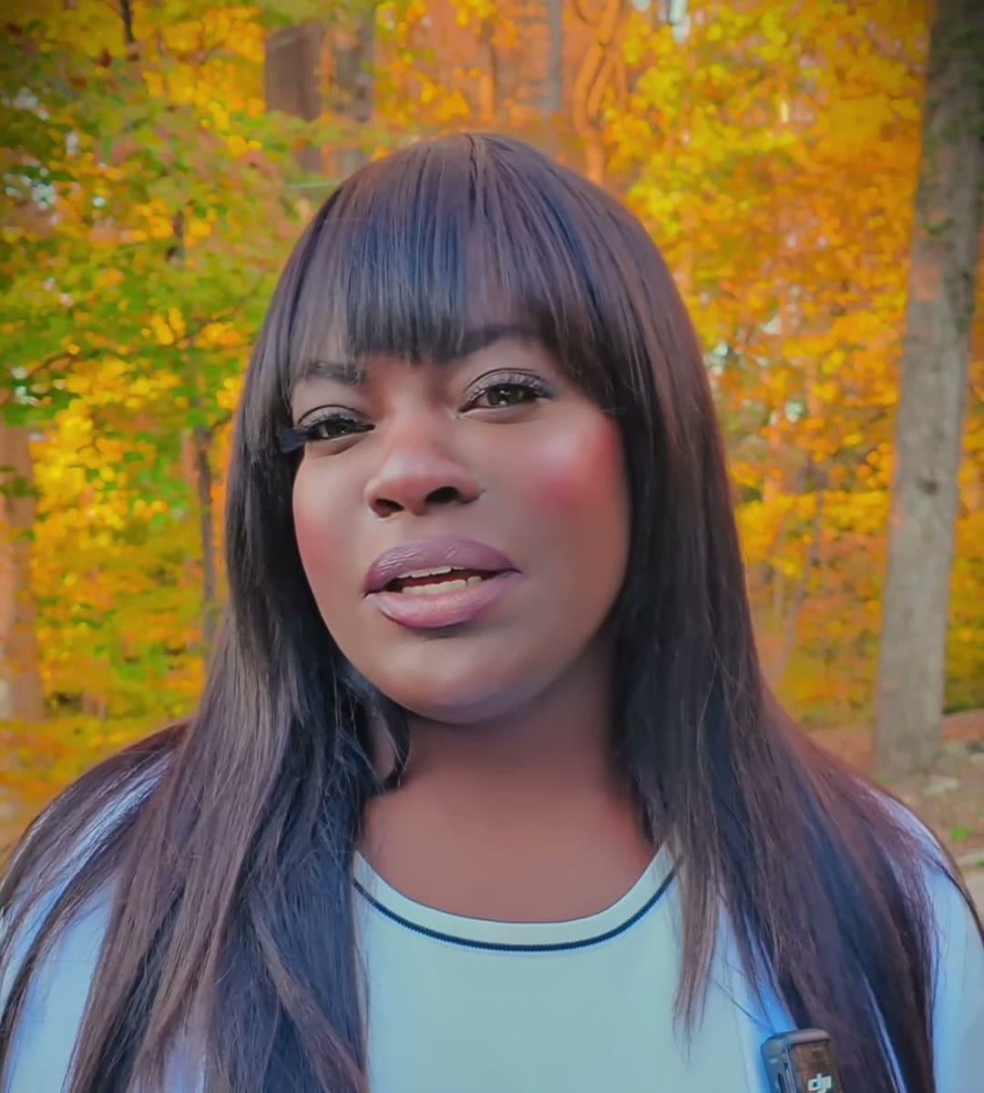 Dr. Raven Baxter, with long dark hair and bangs, speaks to the camera in front of trees with yellow and orange autumn leaves.