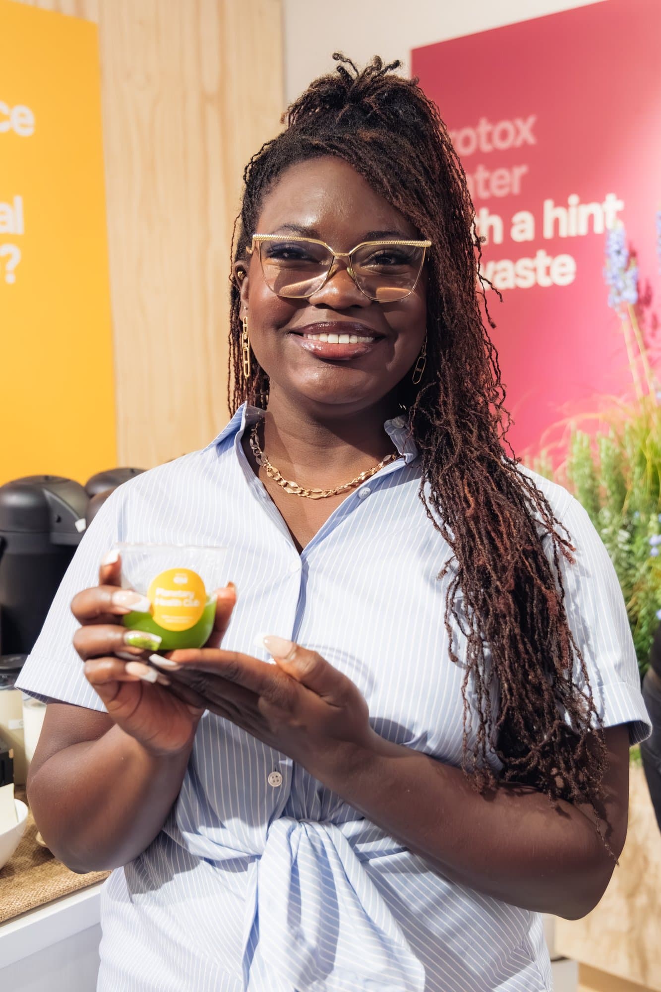 Dr. Raven Baxter, in glasses and a blue striped shirt, smiles at a juice bar while holding a green Planetary Health Club cup.