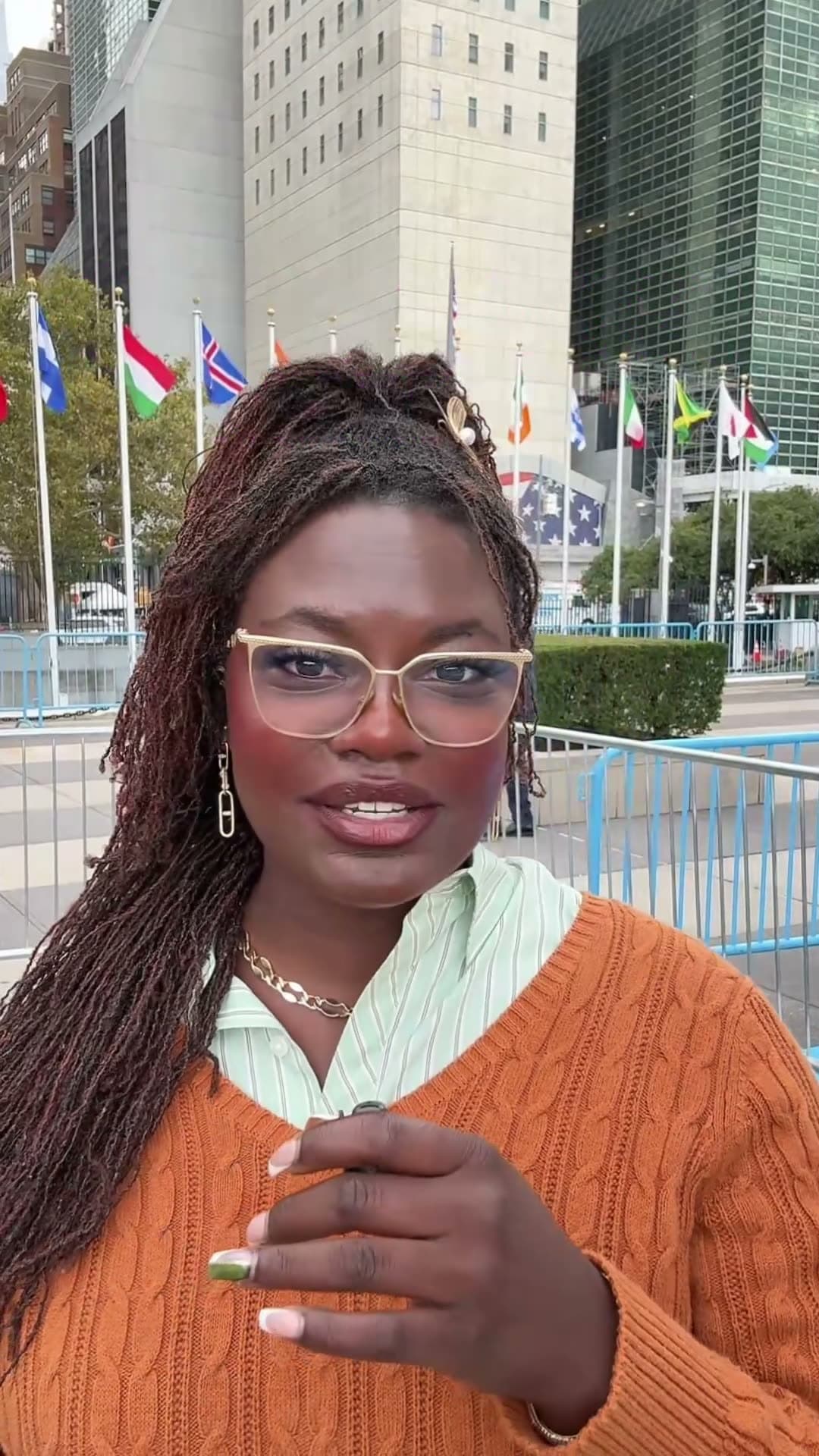 Dr. Raven Baxter, in glasses, an orange cable-knit sweater and a striped collared shirt, speaks to the camera in front of a row of national flags outside United Nations headquarters.
