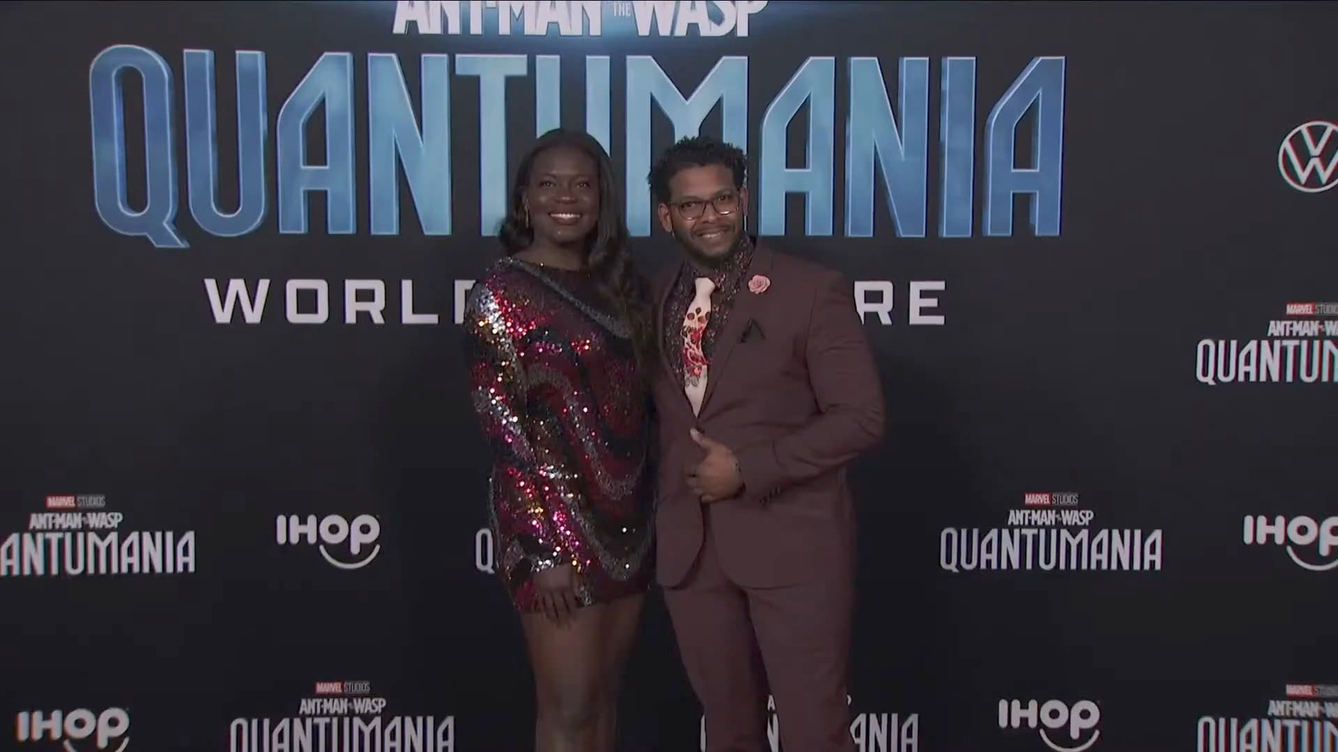 Dr. Raven Baxter, in a multicolored sequined minidress, smiles beside a man in a maroon suit in front of the Quantumania World Premiere press wall.