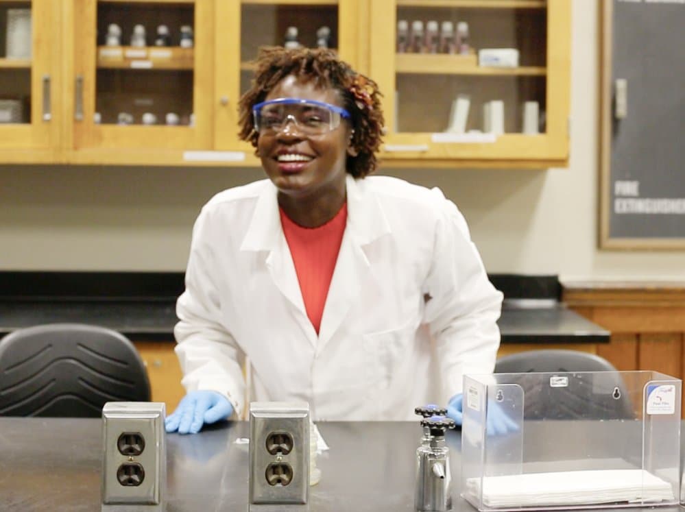 Raven Baxter, in a white lab coat, safety goggles and blue gloves, smiles at a black lab bench in front of wooden supply cabinets.