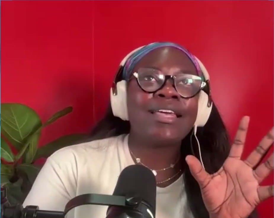 Dr. Raven Baxter, in white headphones, glasses and a white T-shirt, raises one hand as she speaks into a microphone in front of a red wall.
