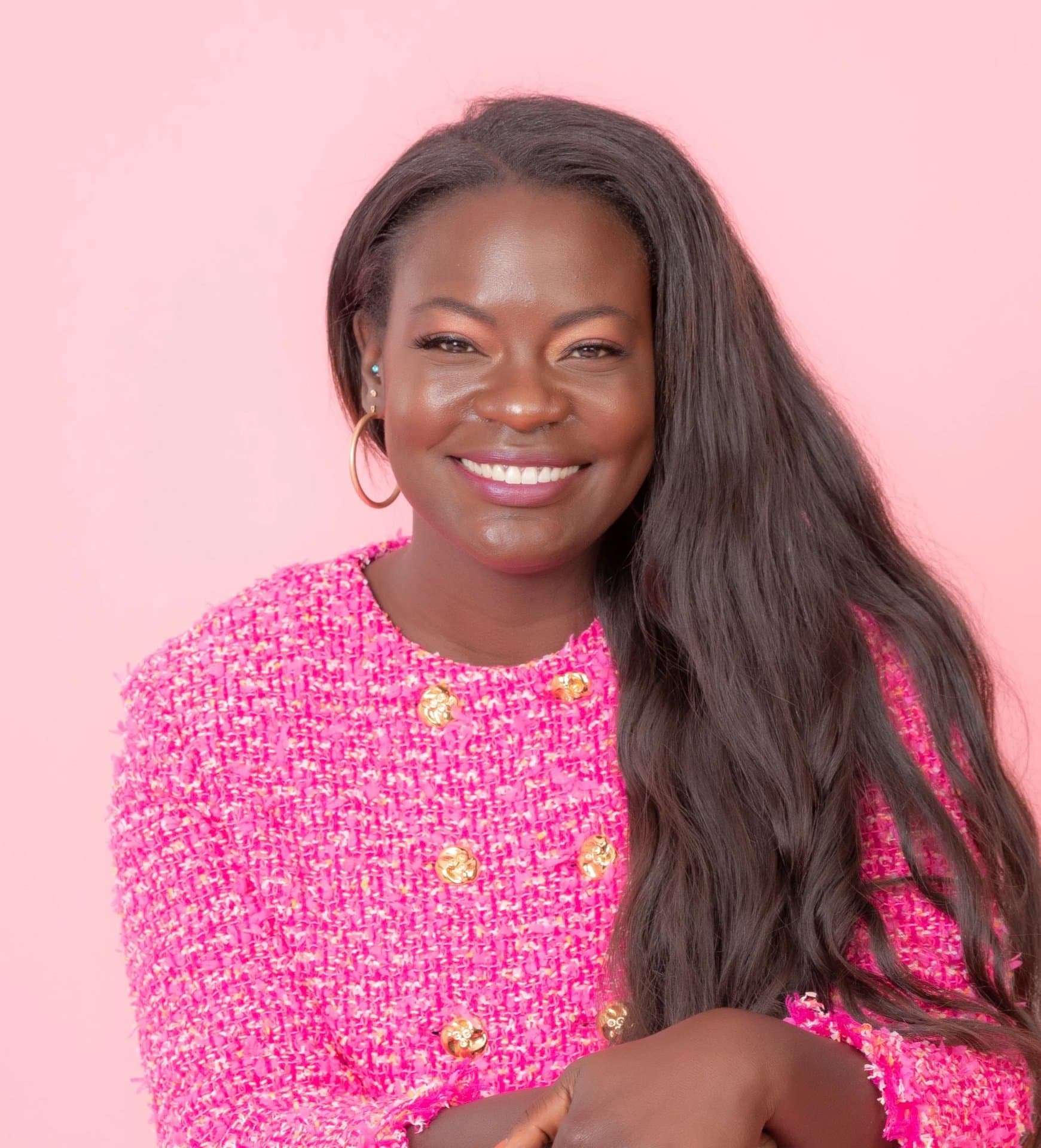Dr. Raven Baxter smiles at the camera in a pink tweed jacket with gold buttons, long dark hair over one shoulder, against a pink wall
