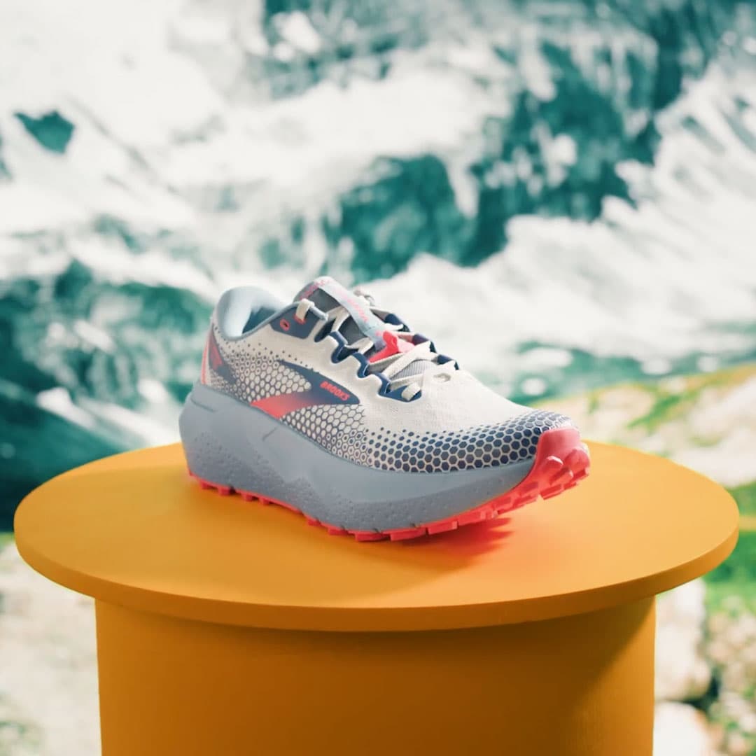 A Brooks Caldera trail shoe on a mustard pedestal in front of a mountain backdrop