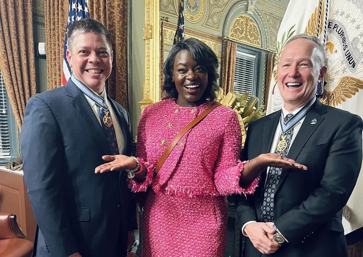 Dr. Raven Baxter in a pink tweed suit, laughing between the two honorees at the Congressional Space Medal of Honor ceremony at the White House, both men wearing their medals