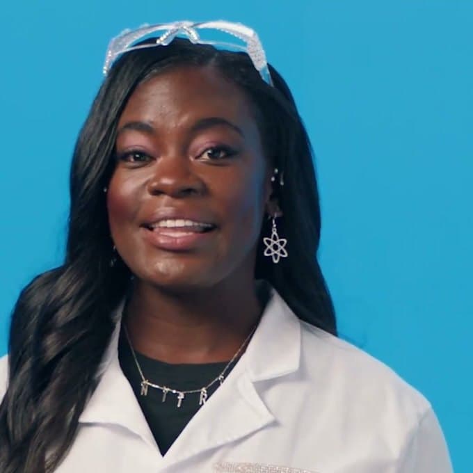 Dr. Raven Baxter in a lab coat with safety glasses pushed up, on bright blue