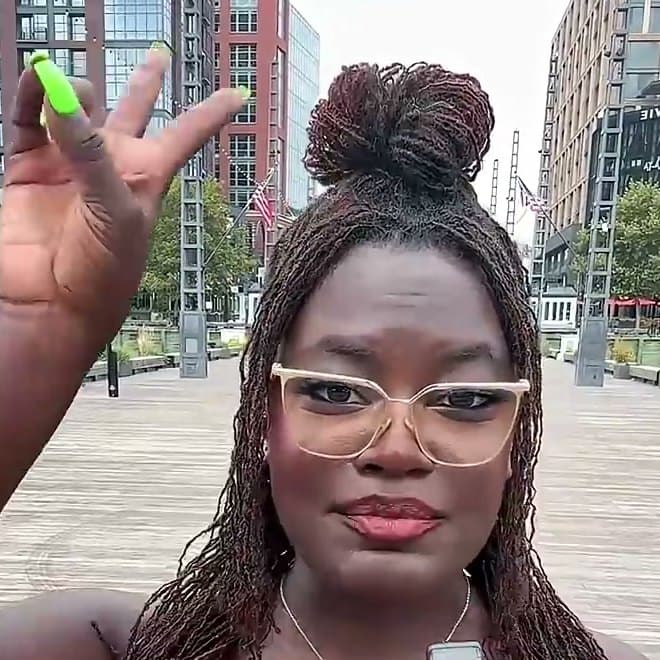 Dr. Raven Baxter in clear glasses, her hand raised to the camera on a city waterfront