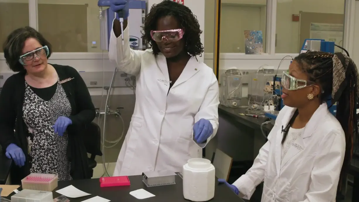 Dr. Raven Baxter in a lab coat and pink goggles holding up a pipette at the bench with Dr. Sarah Bowman and Sydney Anderson, from Nerdy Jobs Episode 1