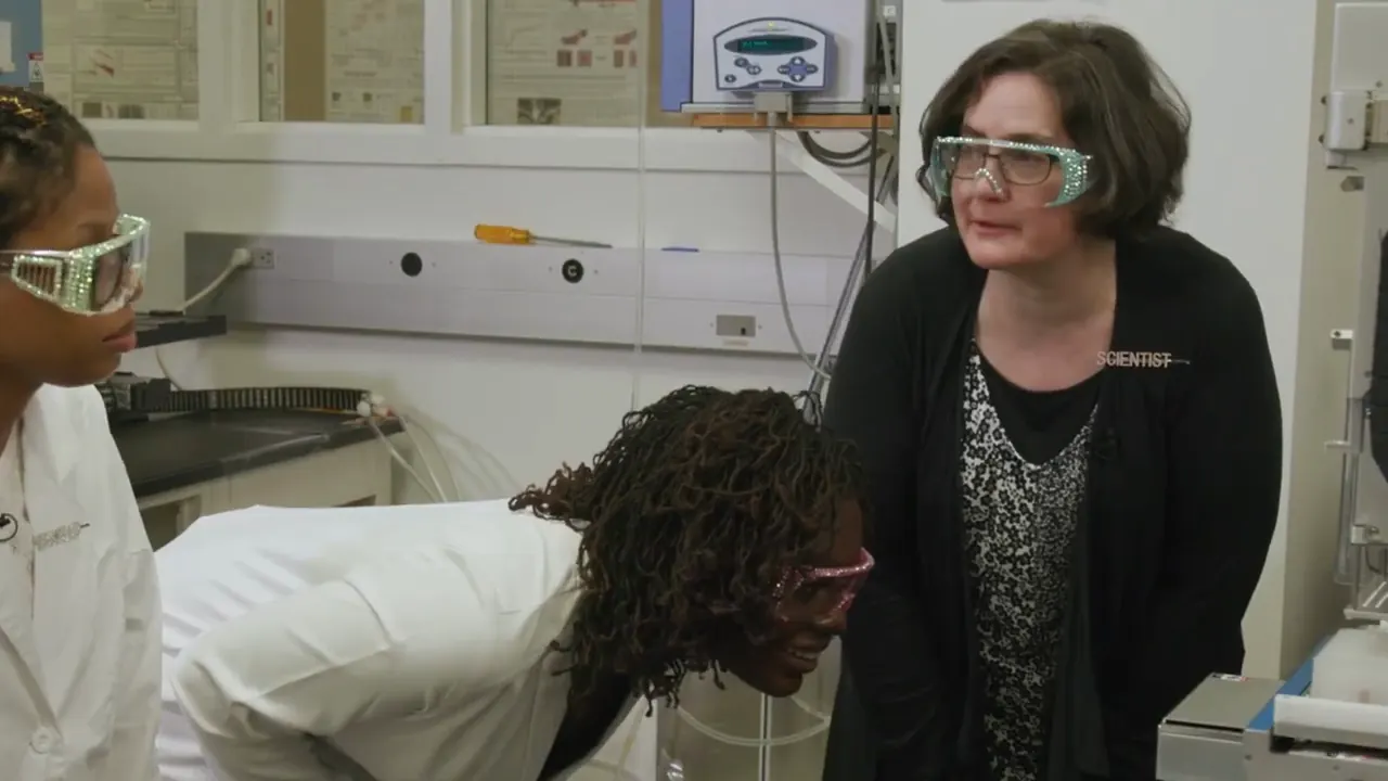 Dr. Raven Baxter in a lab coat and pink goggles bending down to look into a lab instrument as Dr. Sarah Bowman watches, from Nerdy Jobs Episode 1