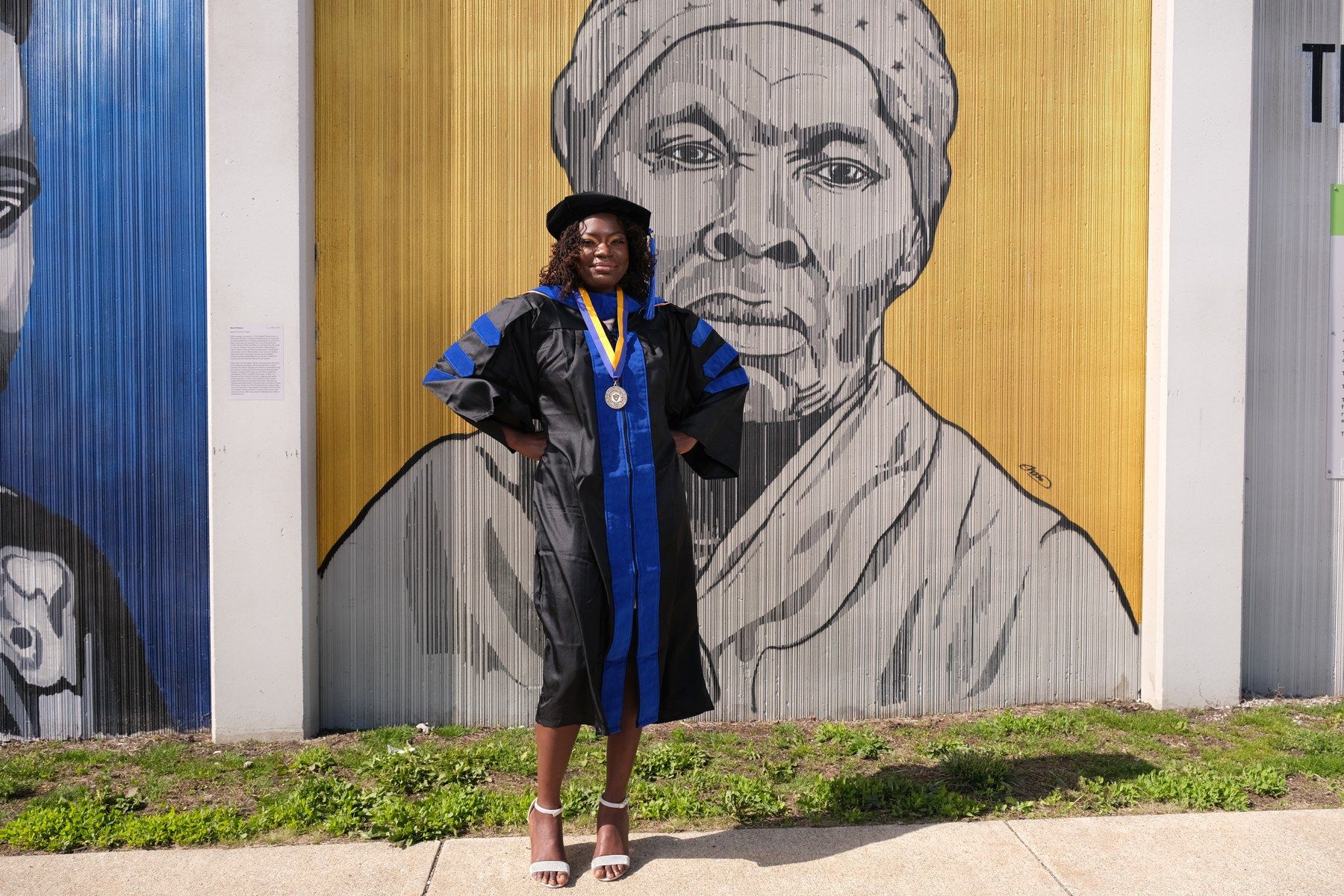 Dr. Raven Baxter in doctoral regalia in front of a Harriet Tubman mural
