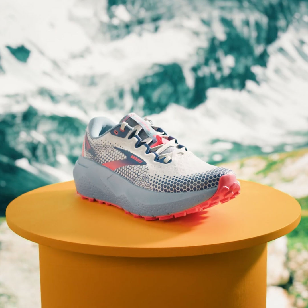 A Brooks Caldera trail shoe on a mustard pedestal in front of a mountain backdrop