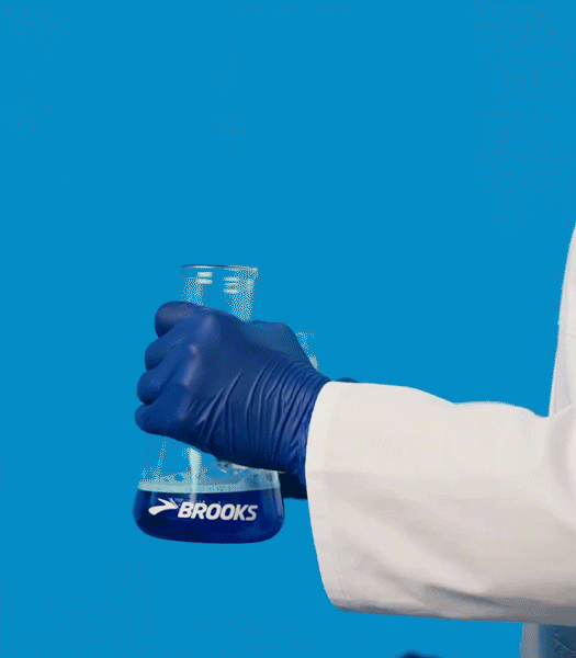 A gloved hand in a lab coat holding a bubbling Brooks-branded Erlenmeyer flask